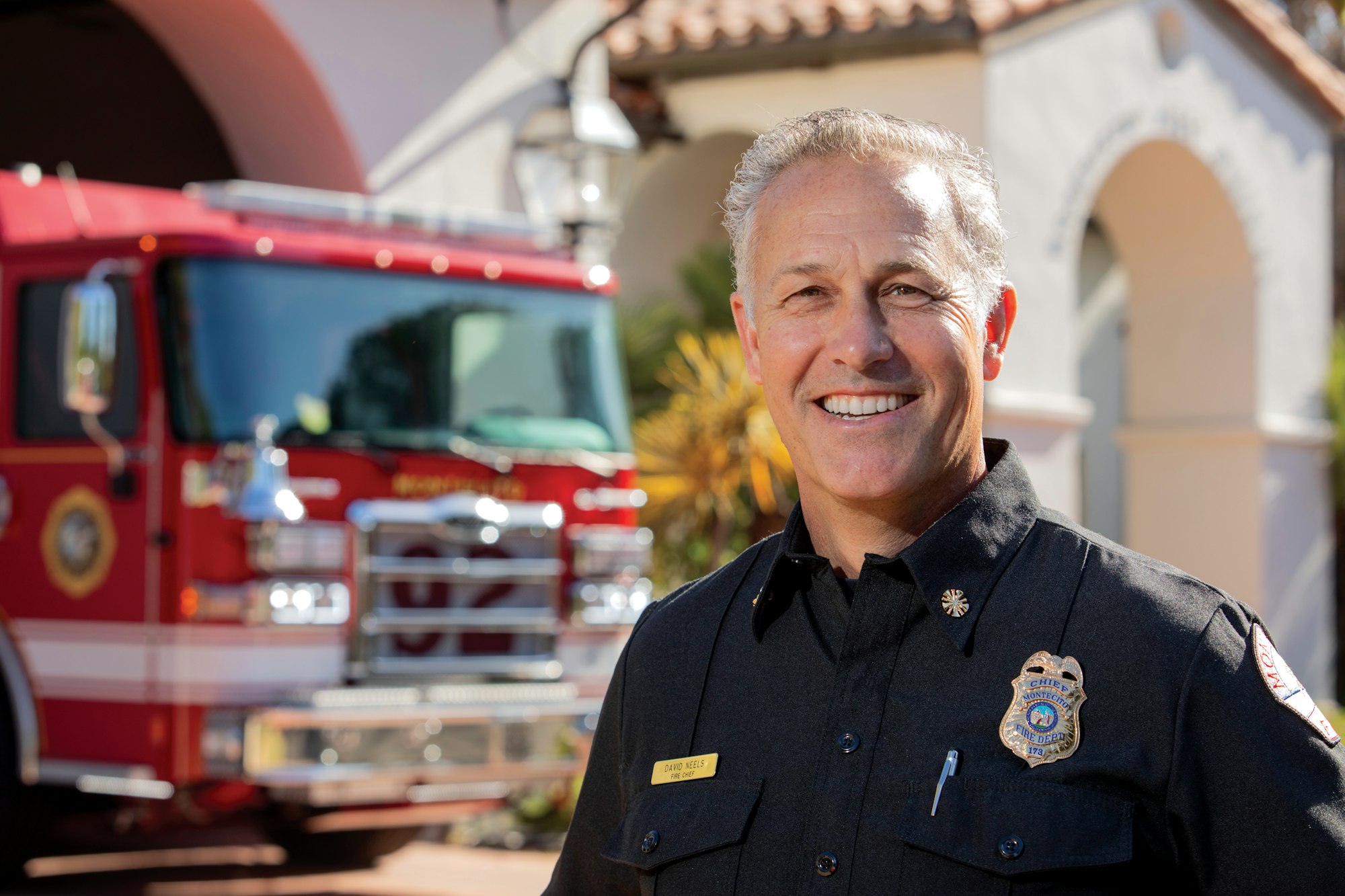 A firefighter in uniform smiling in front of a red fire truck and a building with arches.