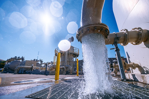 Water is flowing from a large pipe onto a grate, with industrial structures in the background and sunlight shining.