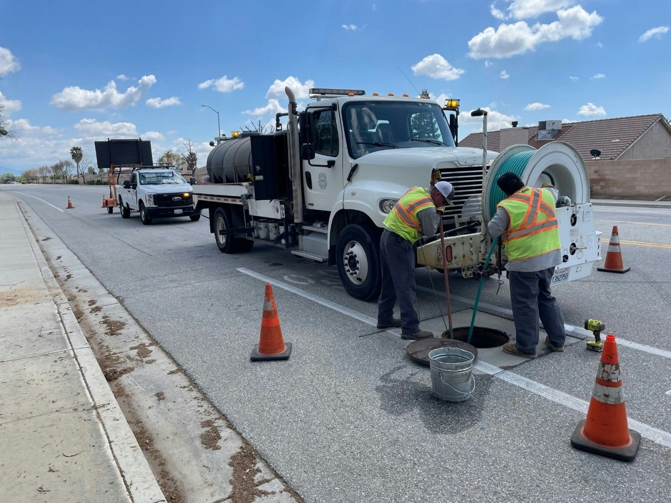 Workers in safety vests performing maintenance near an open manhole, with service trucks and traffic cones on a road.