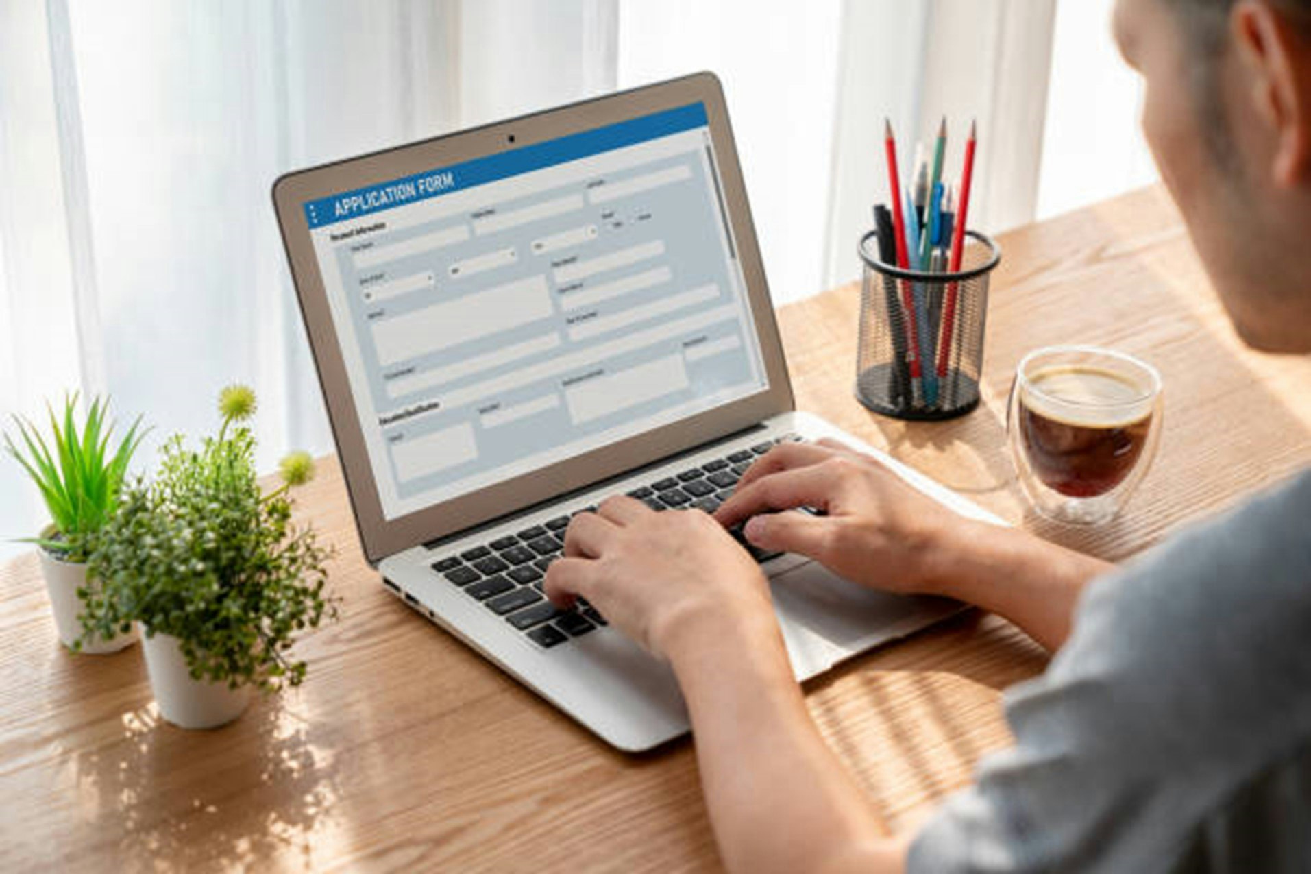 A person is filling out an application form on a laptop, with plants, a cup of coffee, and a container of pens nearby.
