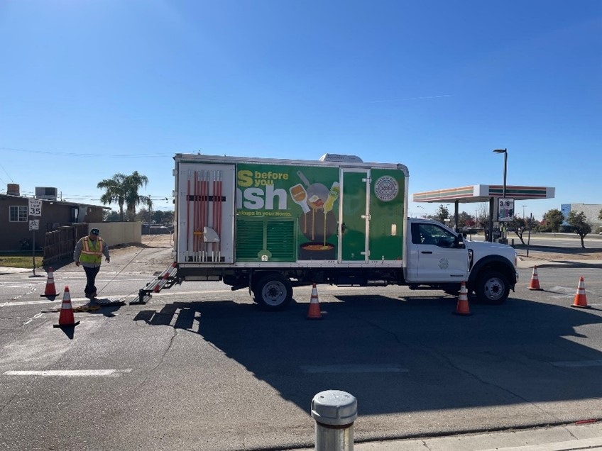 A truck with a green advertisement is parked on the street, surrounded by traffic cones. A person in a safety vest is nearby.