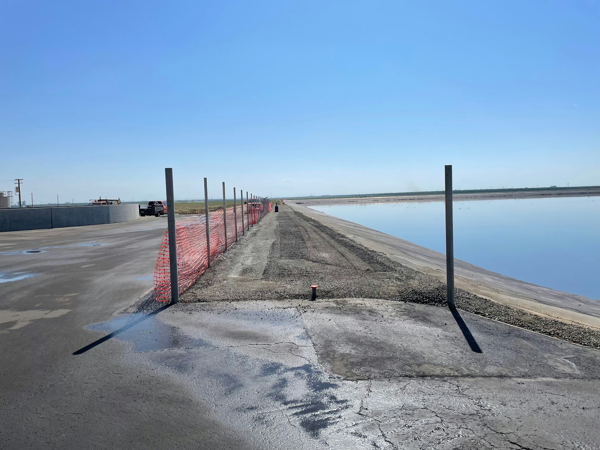 Construction site with dirt, red fencing, and vehicles near a body of water under a clear blue sky.