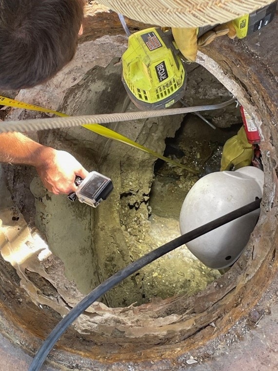 A person inspecting a hole with a camera, a helmet, and a flashlight inside the opening. Tools and equipment are visible.