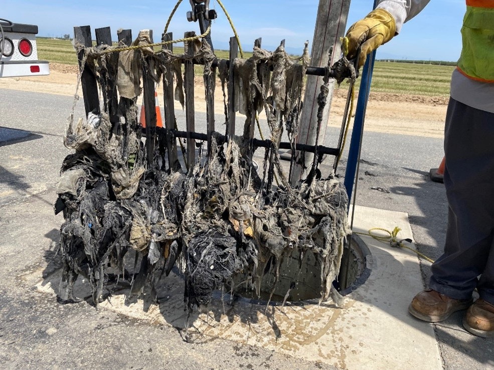 A large mass of wet wipes and debris tangled on a steel grate above a manhole, with a worker in the background.