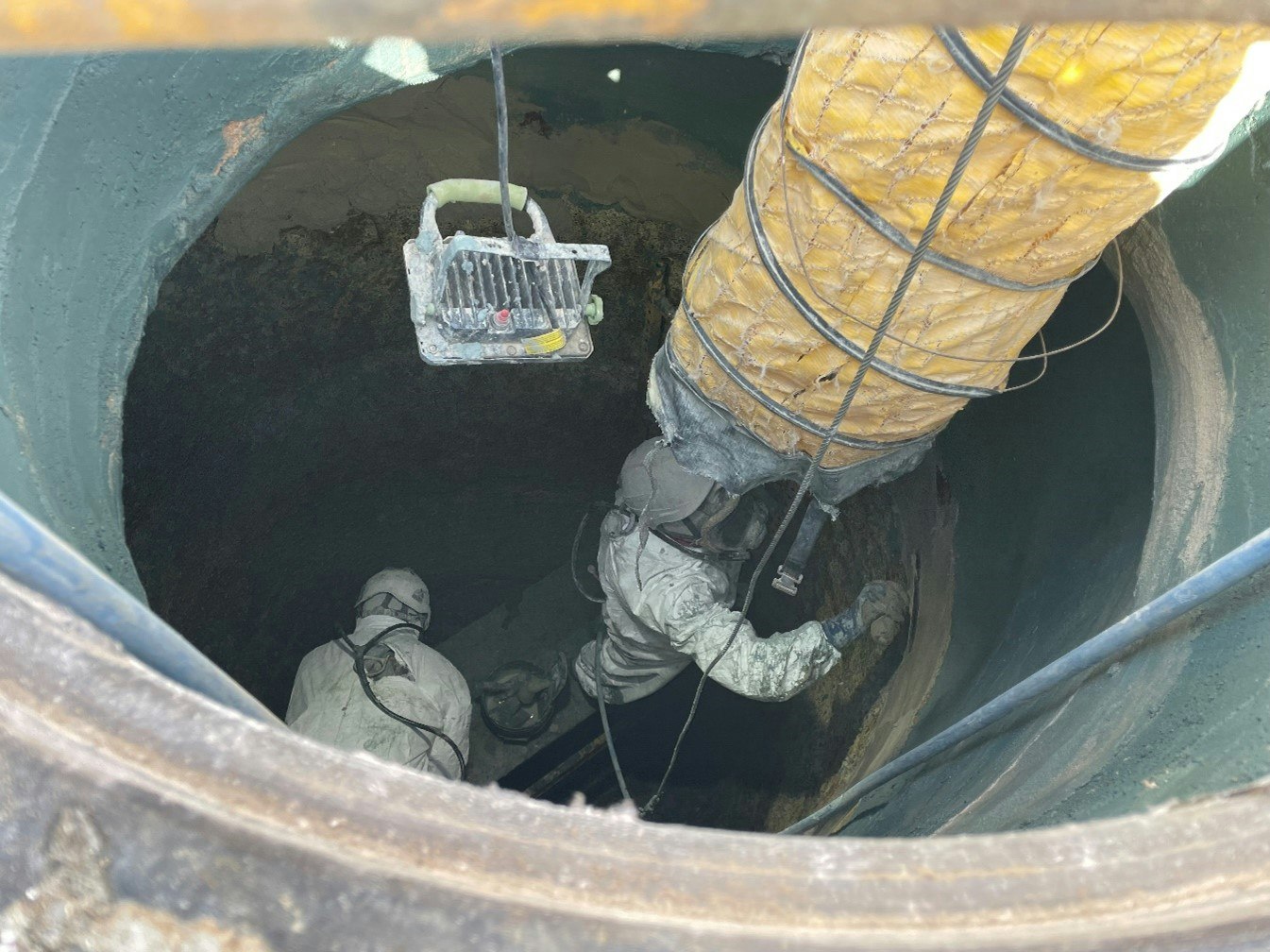 Two workers in protective suits are inside a large pipe or tank, equipped with a light and ventilation duct.
