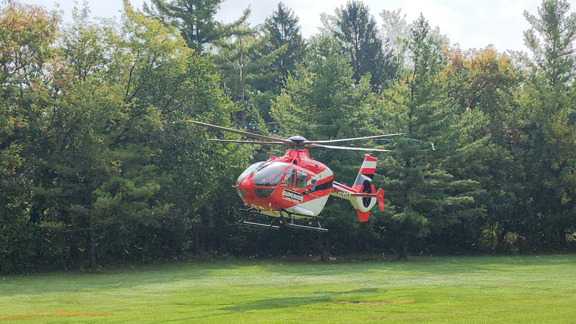 A red and white helicopter hovers above a grassy area, with trees in the background.