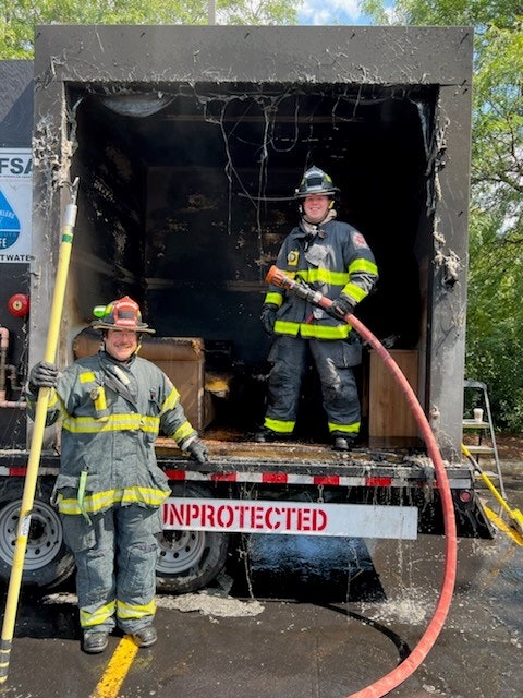Two firefighters in gear, one holding a hose, stand in a partially burnt structure with a sign reading "UNPROTECTED."