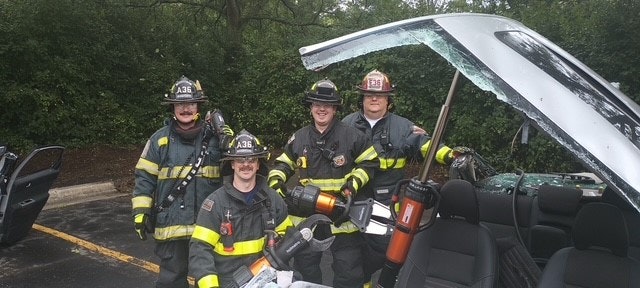 Four firefighters posing with rescue equipment near a car with a removed roof.