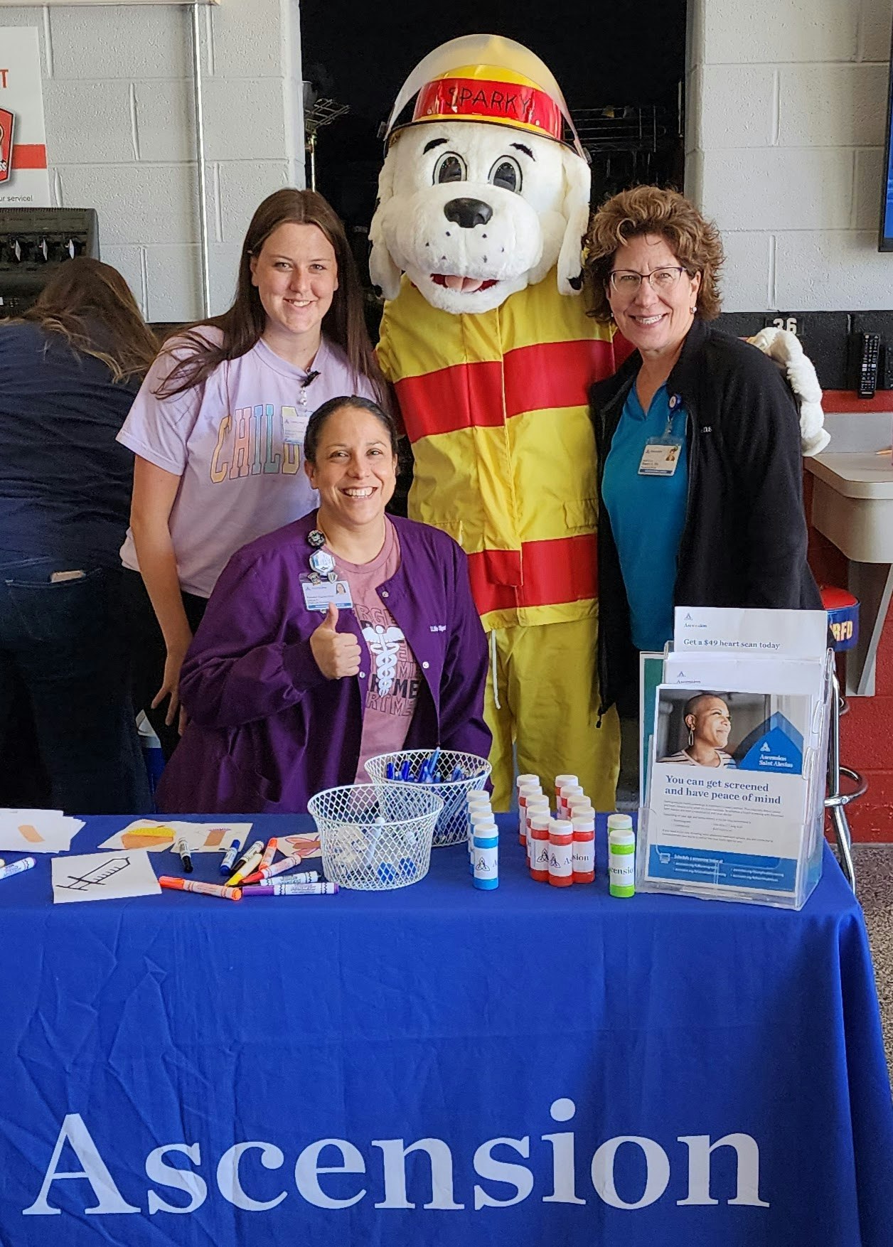 Three people and a costumed character at a table with an Ascension Health banner, markers, and flyers.