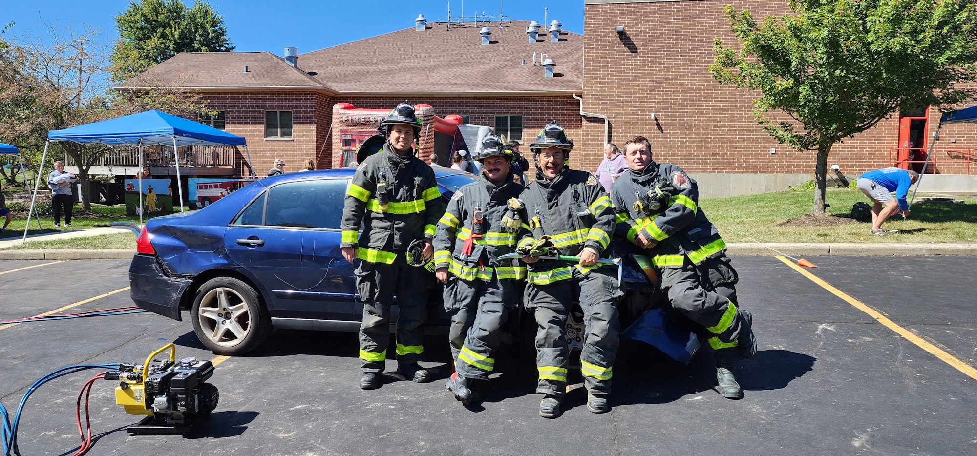Firefighters posing by a damaged car during outdoor event.