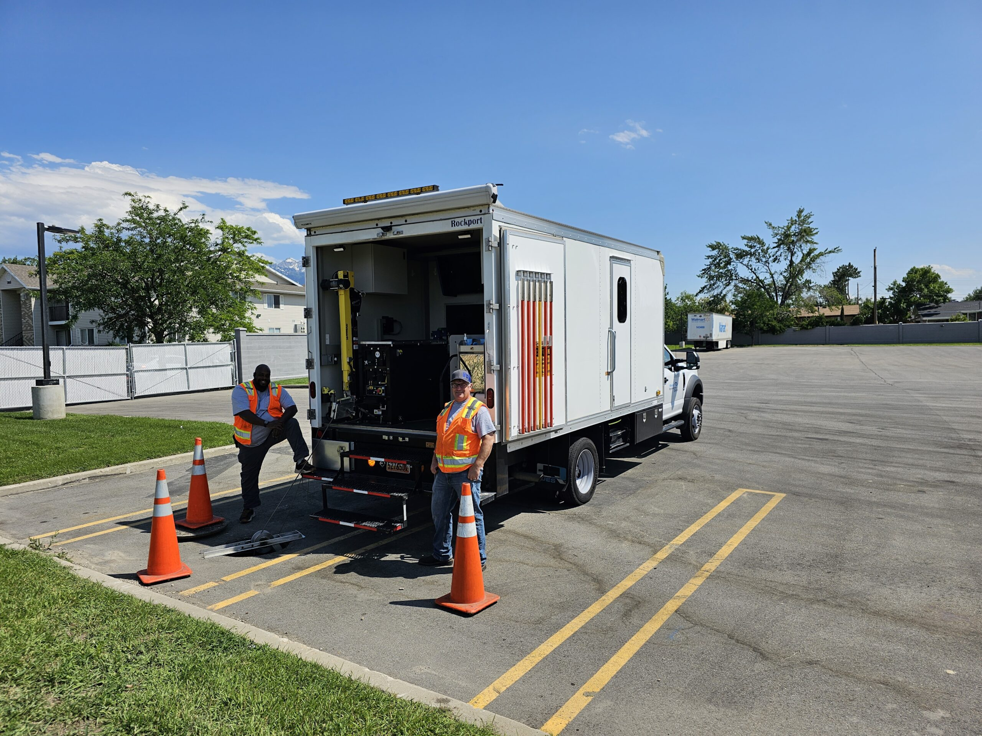 A utility vehicle with an open rear compartment and two workers in safety vests and helmets, surrounded by traffic cones.