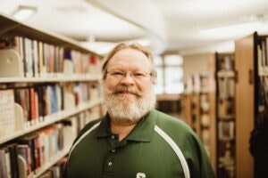 A person with a beard wearing a green shirt stands smiling in a library aisle.