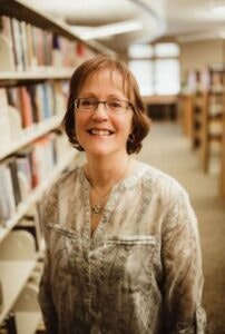 A person smiling in a library, standing between bookshelves.