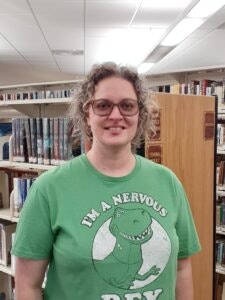 A person with glasses is smiling, standing in a library, wearing a green shirt with a cartoon dinosaur and text.