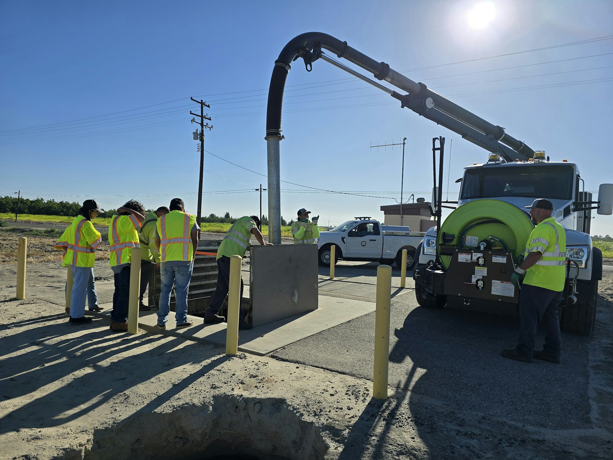 Workers in safety vests operate a truck with a large hose, likely engaged in maintenance or utility work at a site.