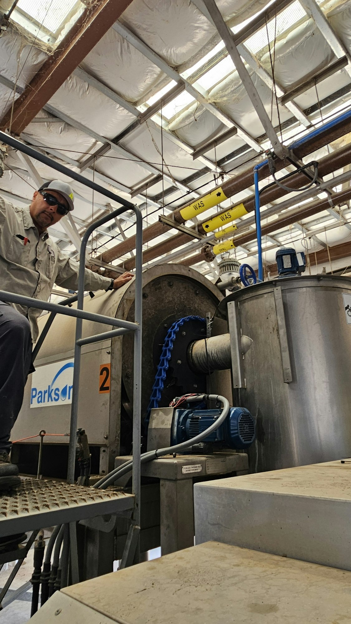 Industrial setting with equipment, labeled pipes, man in hard hat and glasses, and metal walkways.