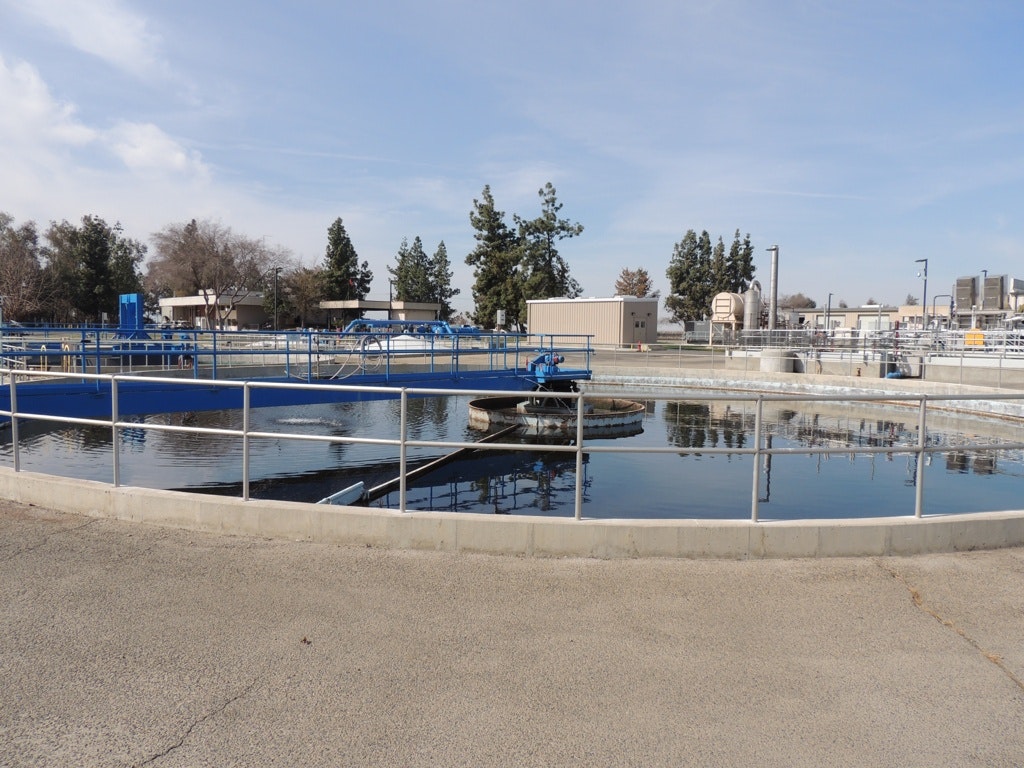 The image shows a water treatment facility with large tanks, machinery, and a fenced area, set against a clear sky.