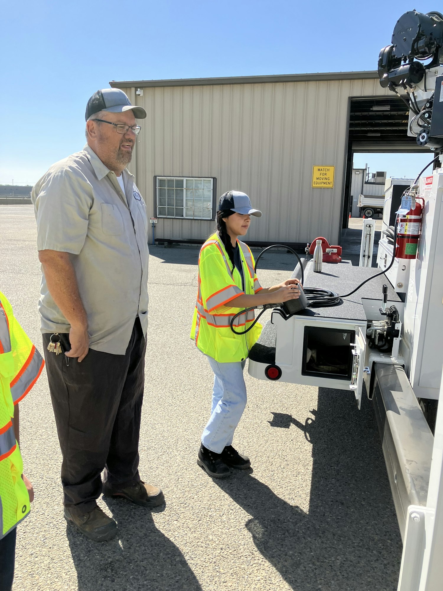 Two people in safety vests and hats operate machinery outdoors near a building.