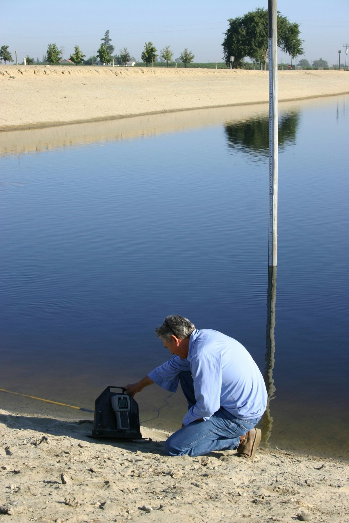 A person kneels by a calm body of water, using equipment on the sandy shore, possibly for environmental testing or measurement.