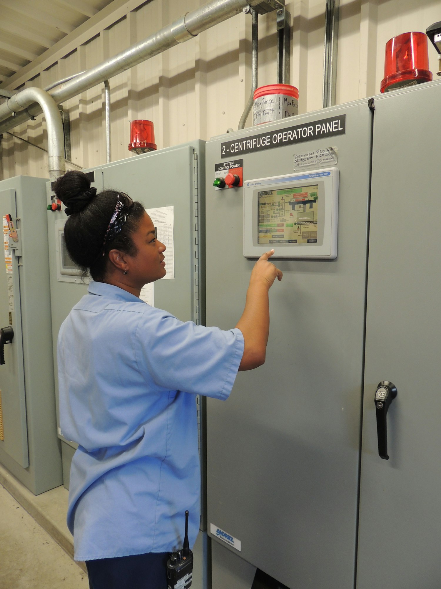 A woman operates a centrifuge control panel in an industrial setting, focusing on a touchscreen display with equipment nearby.