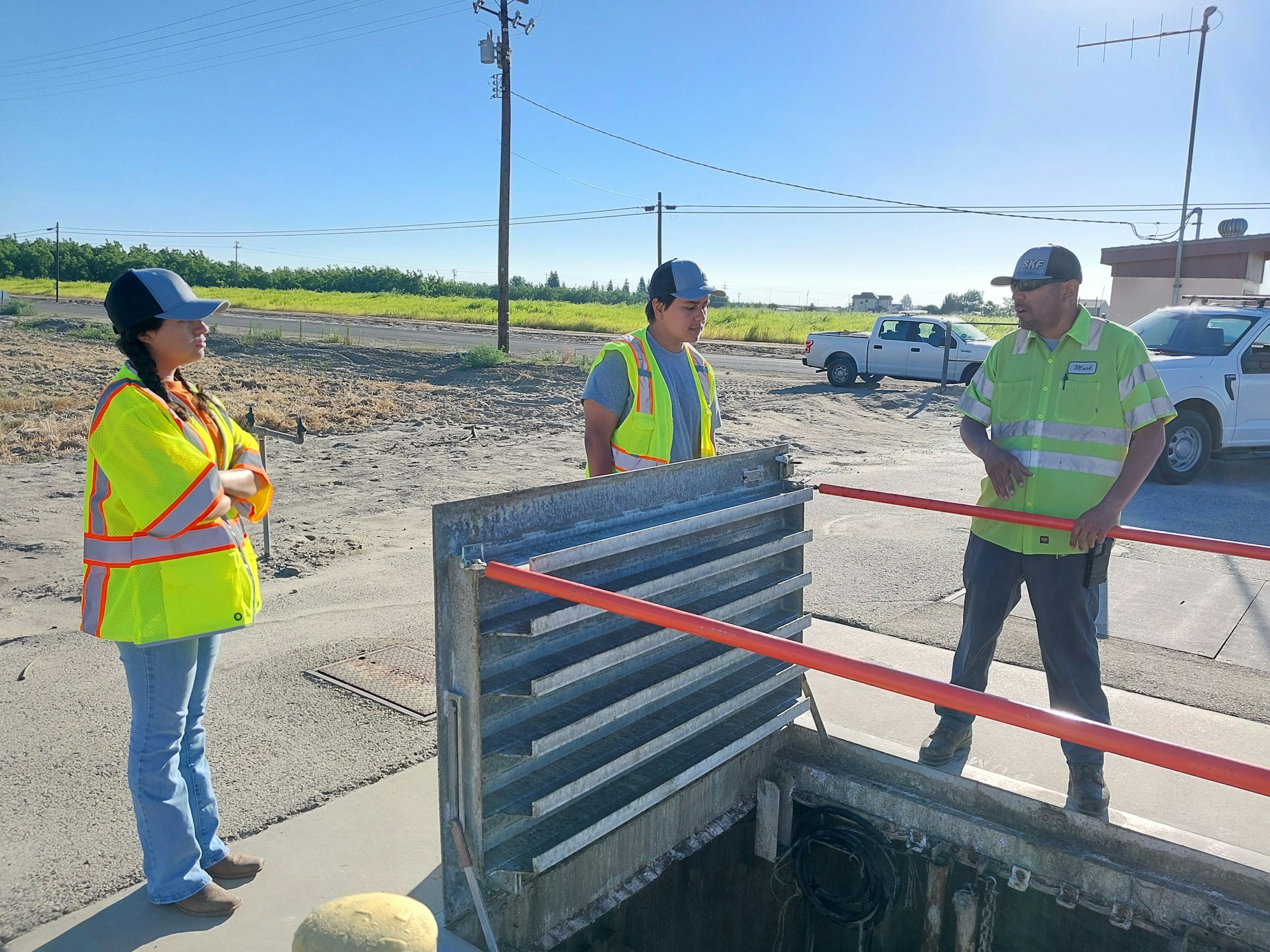 Three people wearing safety vests are gathered around an open utility hole in an outdoor setting with vehicles nearby.