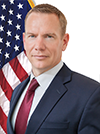 The image features a professional headshot of a person in a suit, standing in front of the American flag.