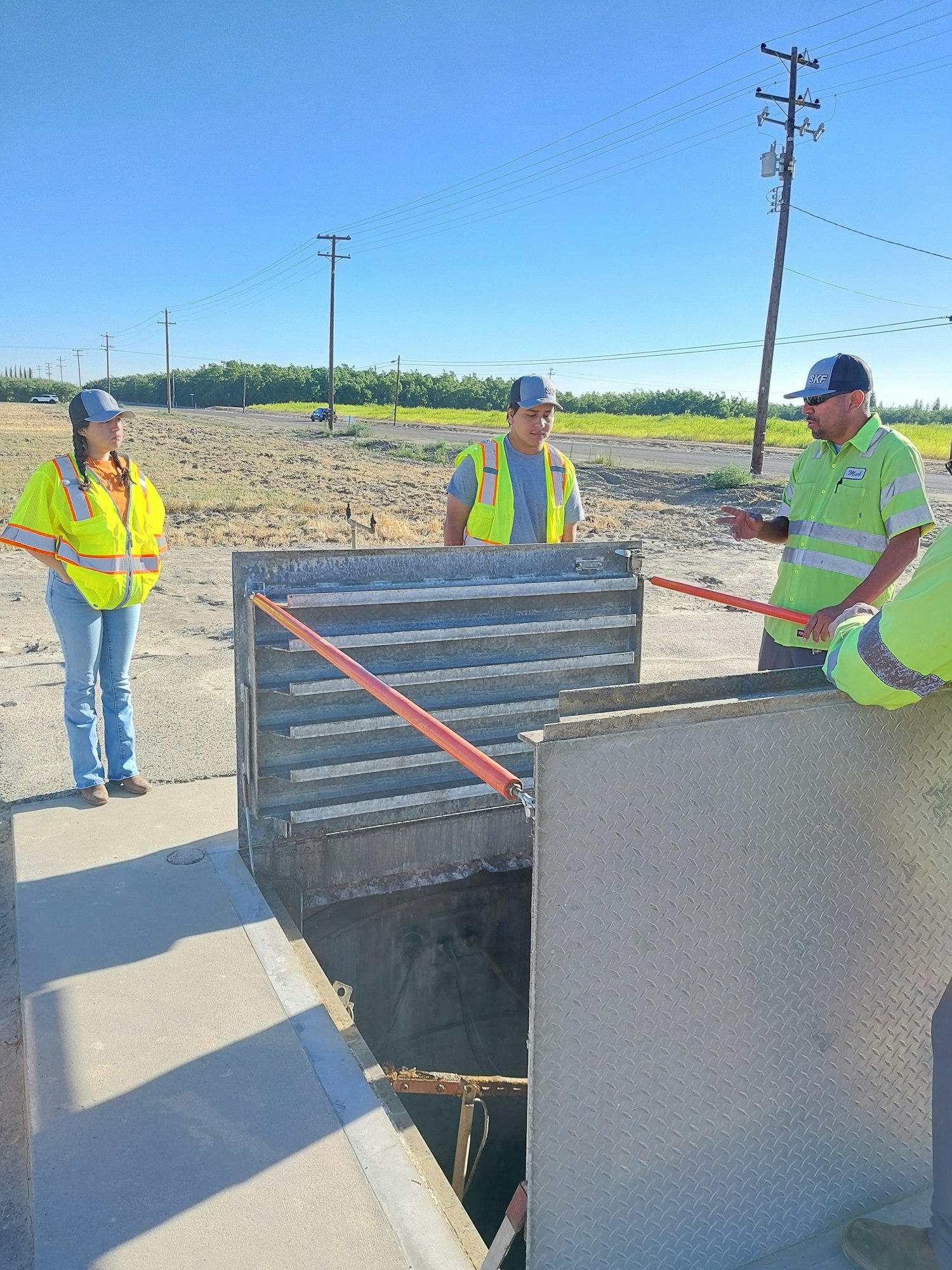 Three people in safety vests standing near an open hatch with a ladder.