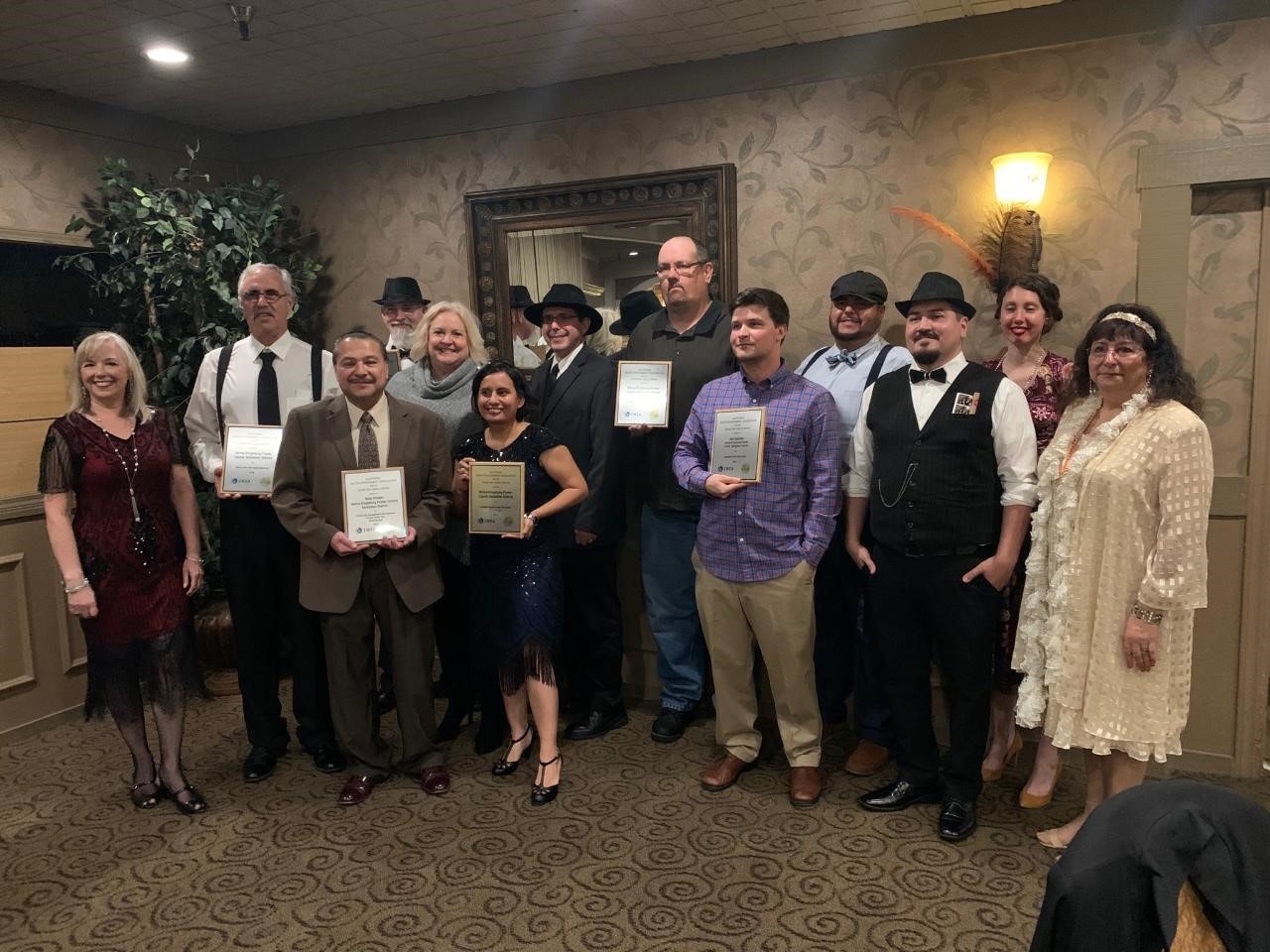 A group of people poses for a photo holding certificates, dressed in formal and vintage attire, in a decorated indoor setting.