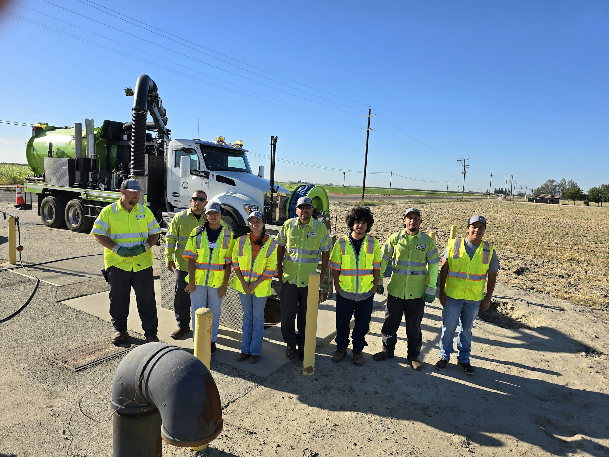 People in safety vests pose in front of a large vehicle with industrial equipment in a rural area.