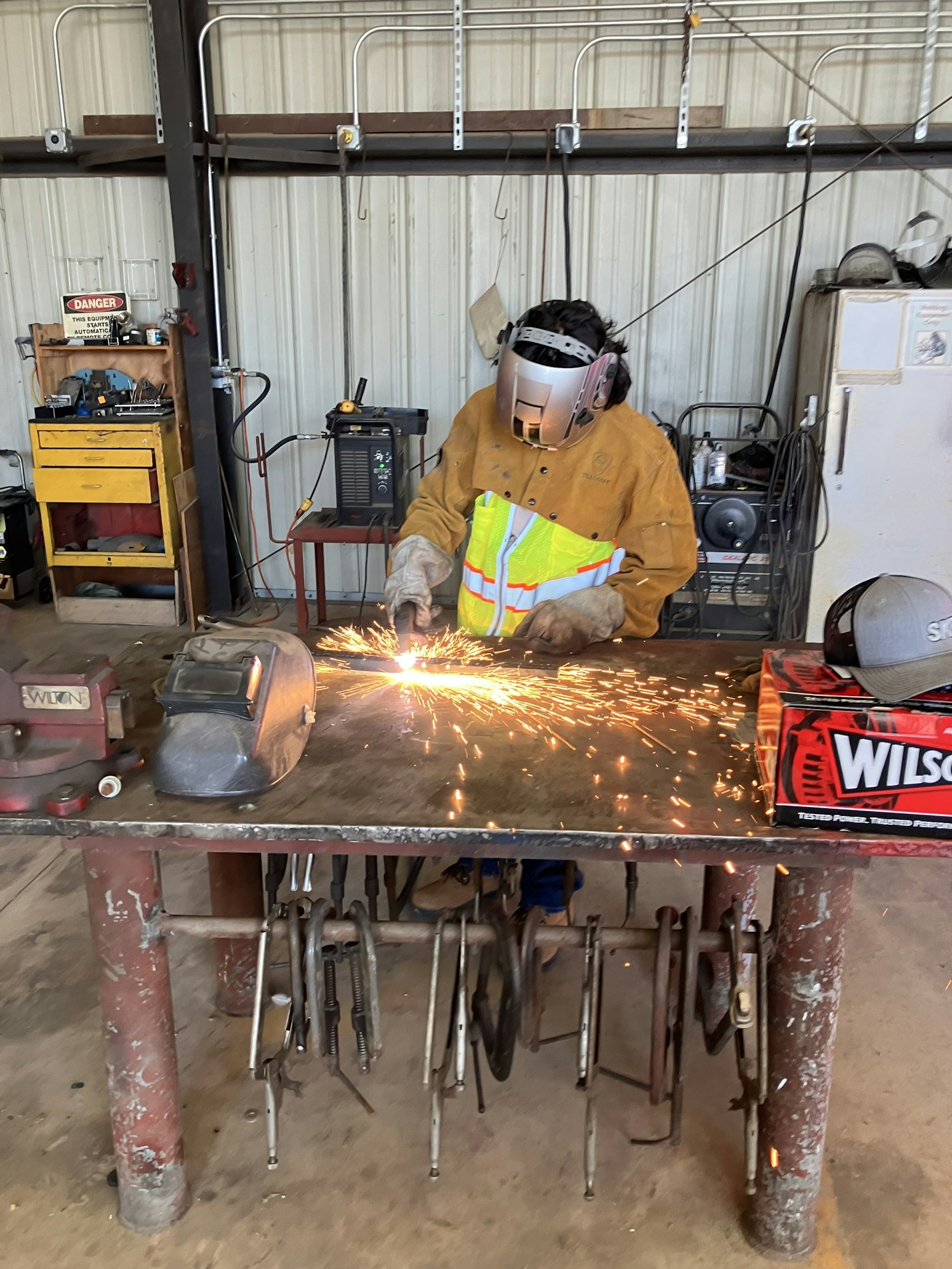 A person is welding in a workshop, wearing protective gear, with sparks flying around.
