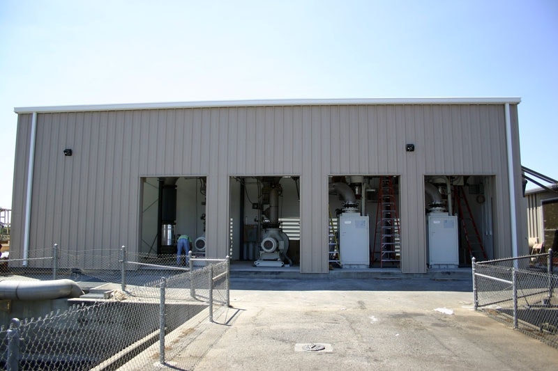The image shows a large metal building with three open bays, some machinery inside, and a fenced area nearby.