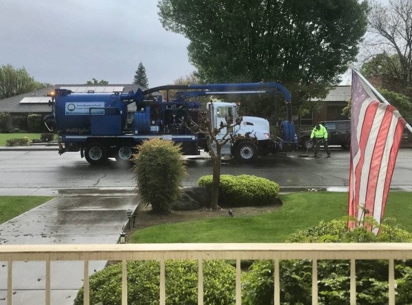 A large blue maintenance truck is parked on a rainy street, with a worker in a yellow jacket nearby and an American flag in the foreground.