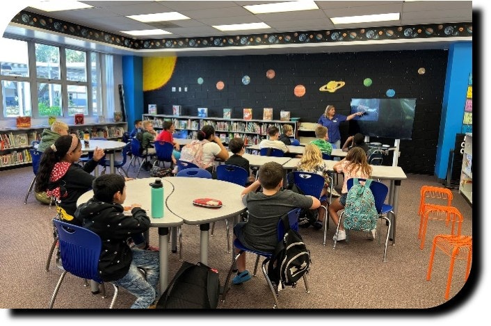 Children sitting at tables in a colorful classroom with a space theme, listening to a teacher by a screen.