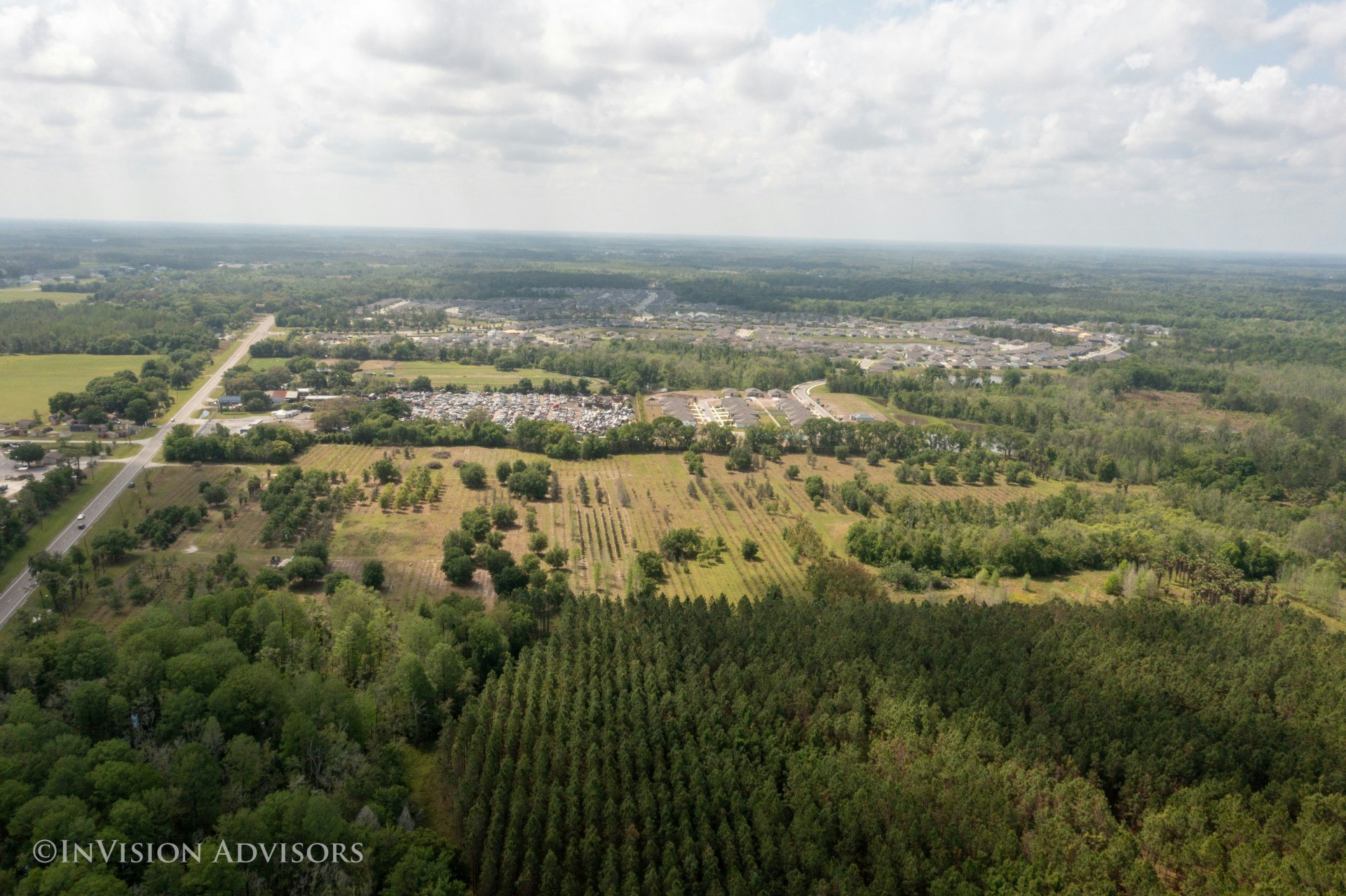 Aerial view of farmland and forest with a distant residential area and road under a cloudy sky.