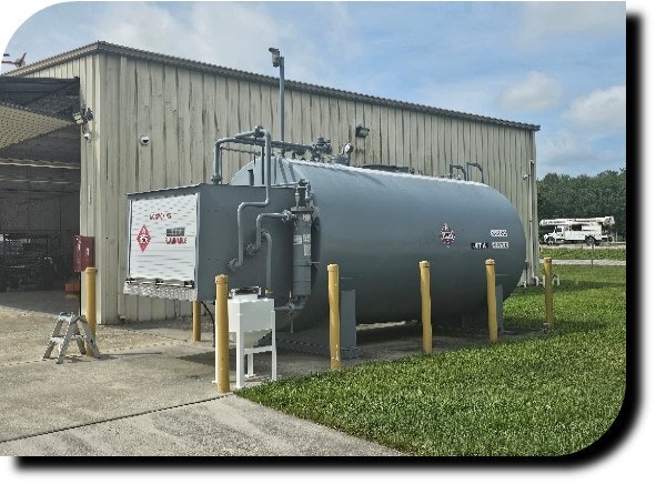 A large industrial tank with safety labels outside a warehouse, protected by yellow bollards.