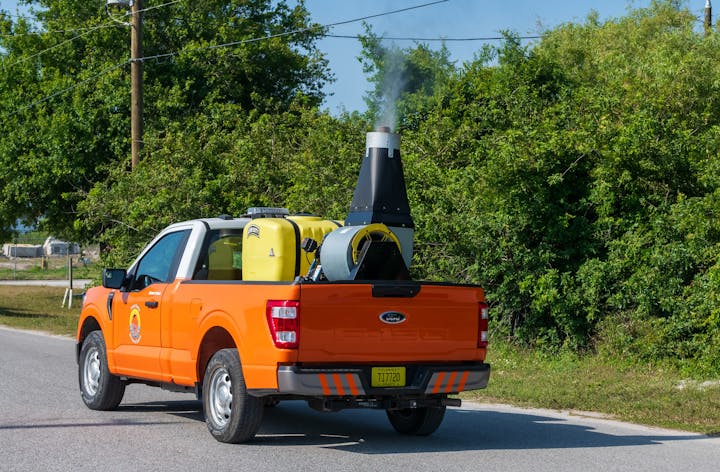 An orange pickup truck with sprayers and equipment, possibly for mosquito control, driving on a road near trees.