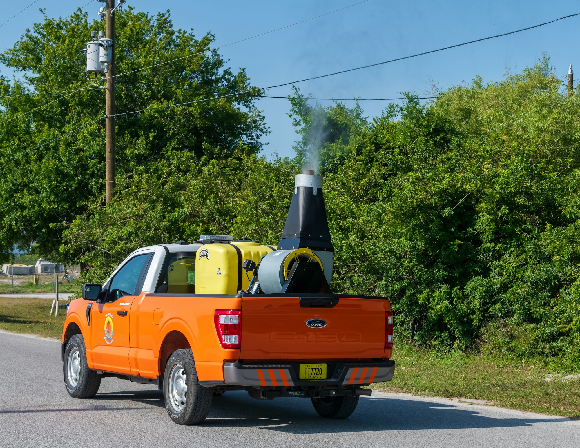 An orange truck with equipment, possibly for fumigation, is on the road emitting smoke.