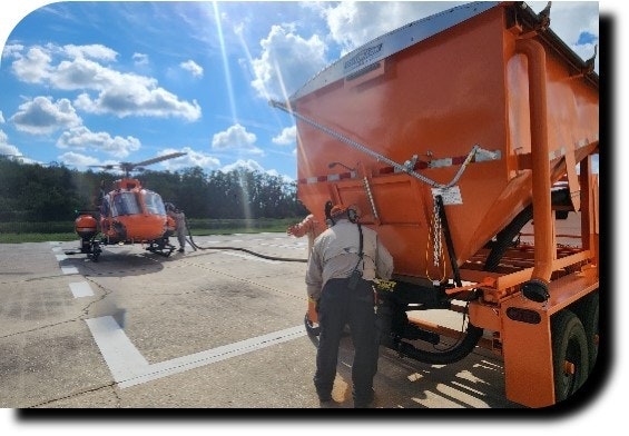A person connects a hose from an orange tanker to a helicopter on an airstrip under a bright sky.