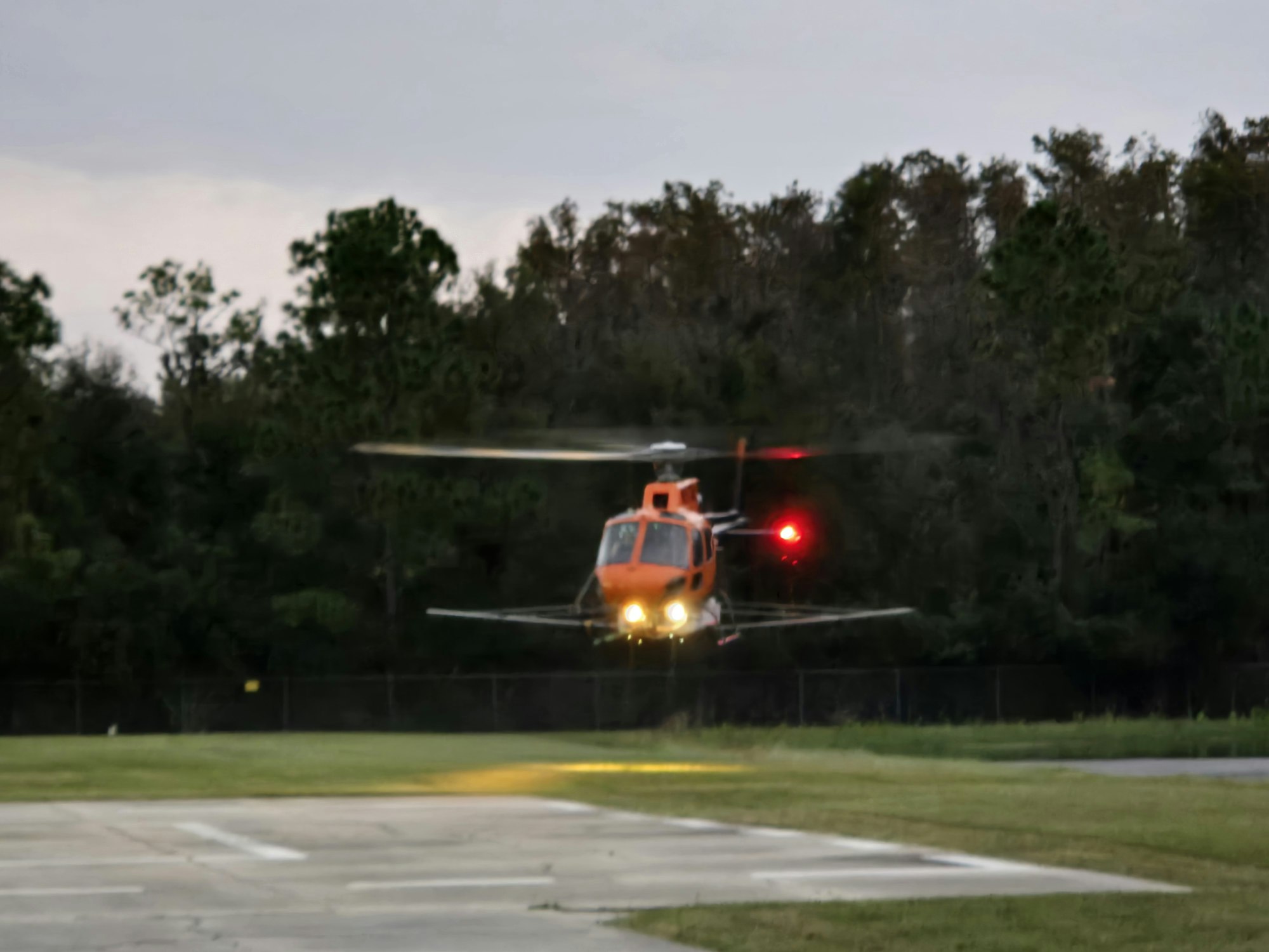 A red and orange helicopter is hovering above a grassy area, near trees, with its lights on.