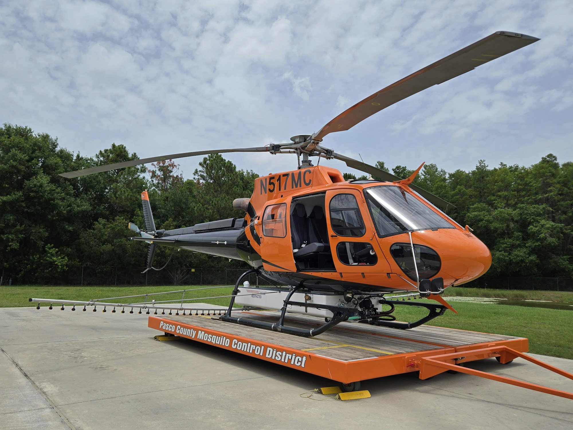 Orange helicopter on a platform labeled "Pasco County Mosquito Control District," surrounded by greenery.