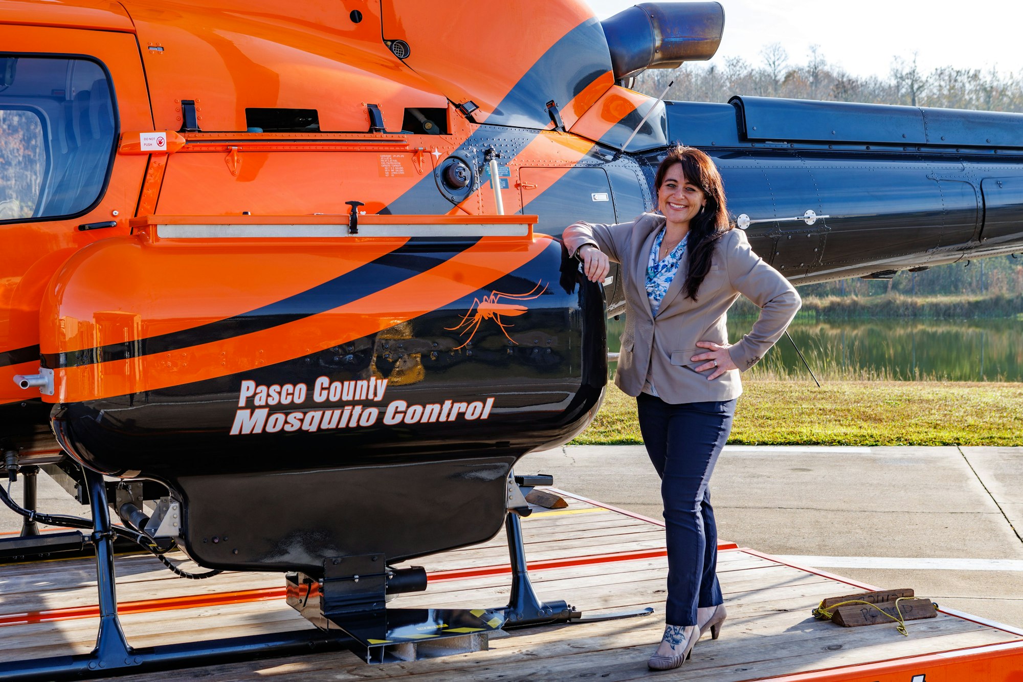 Woman posing by an orange "Pasco County Mosquito Control" helicopter on a sunny day.
