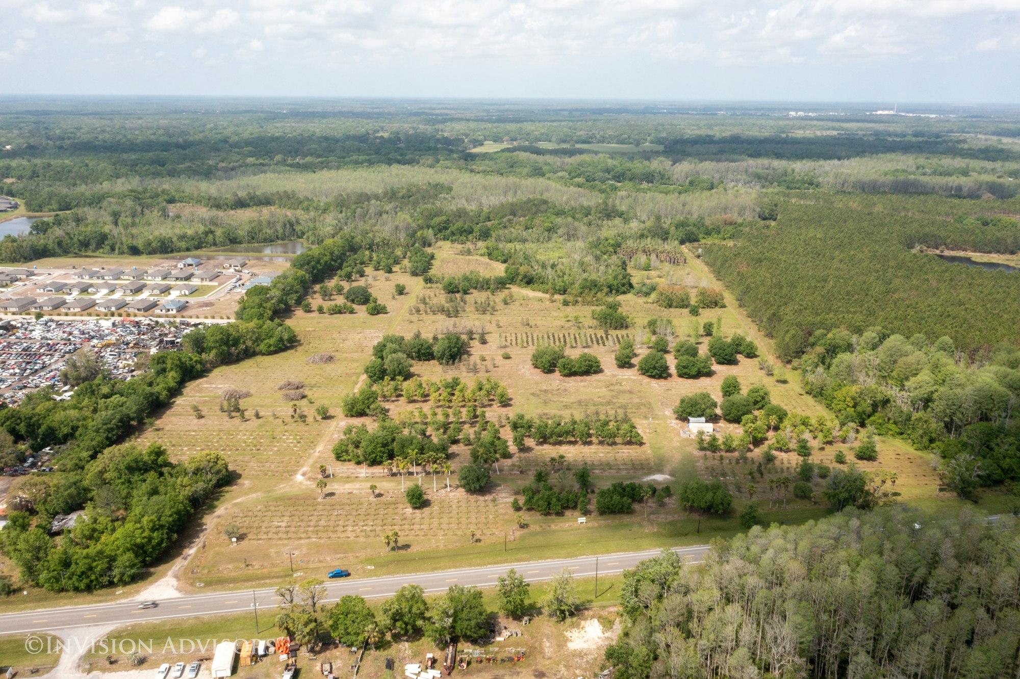 Aerial view of a landscape featuring orchards, wooded areas, a road, and nearby residential structures and a parking lot.