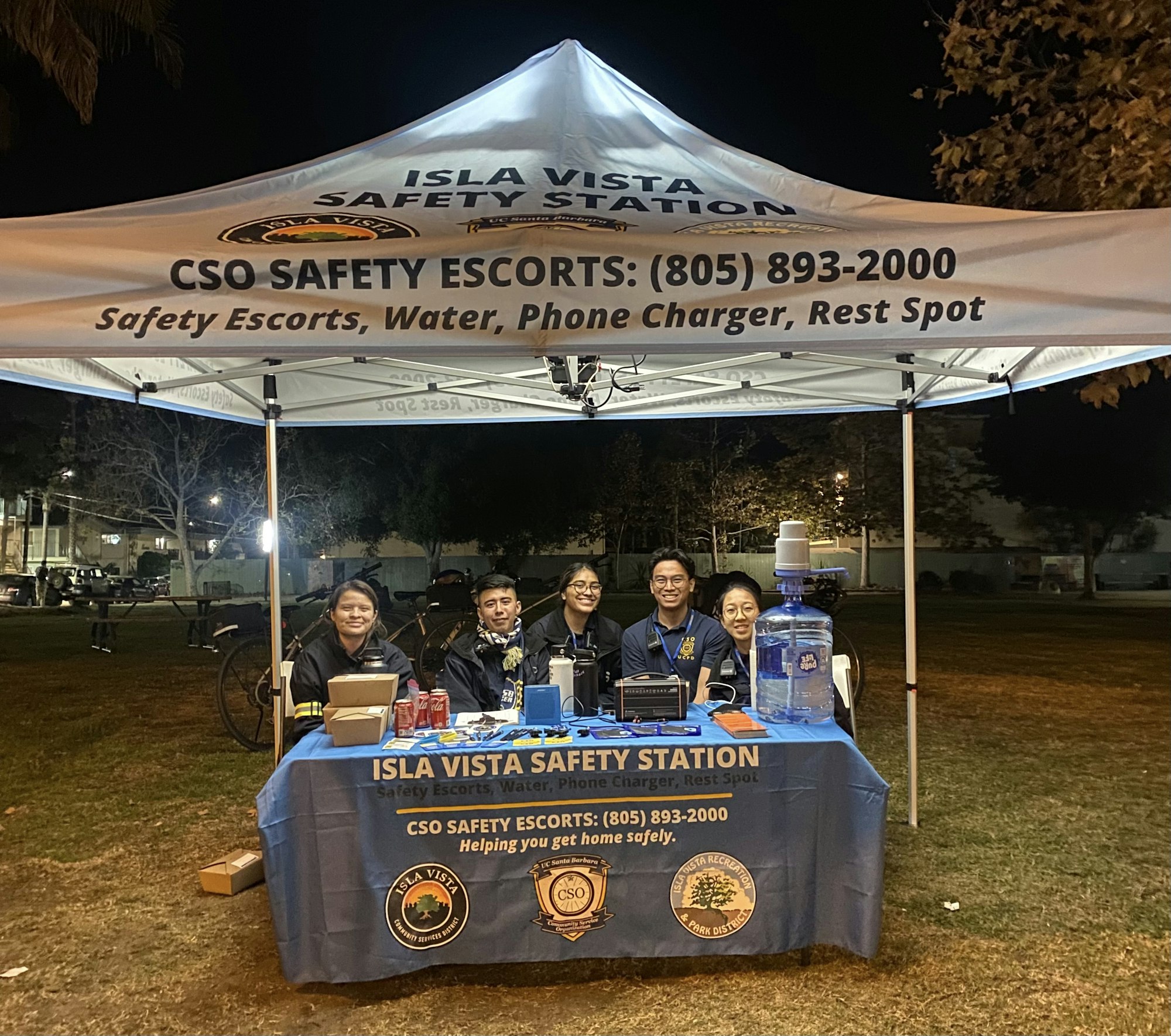 A group of smiling people at a safety station offering escorts, water, phone charging, and a rest spot at night.