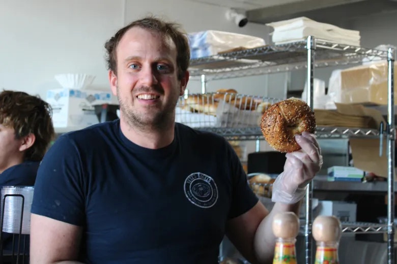 A smiling person holds a bagel in a kitchen or bakery setting, wearing gloves and surrounded by food items.