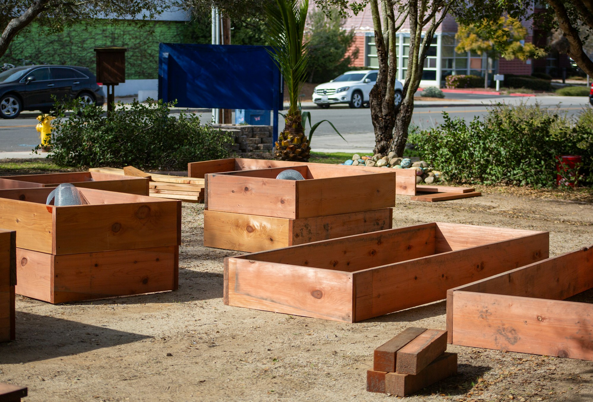 Wooden garden beds being assembled in an outdoor area with trees and cars in the background.