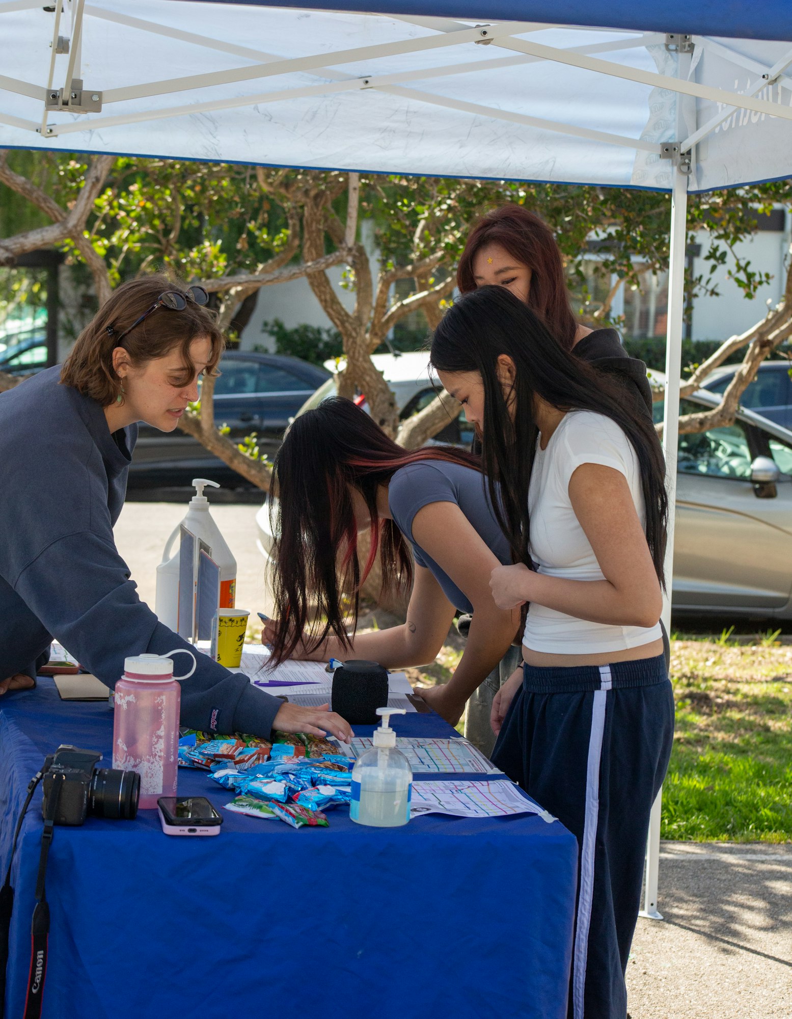 People gathered around a table under a tent, looking at documents, with snacks, hand sanitizer, a camera, and a pink bottle on the table.