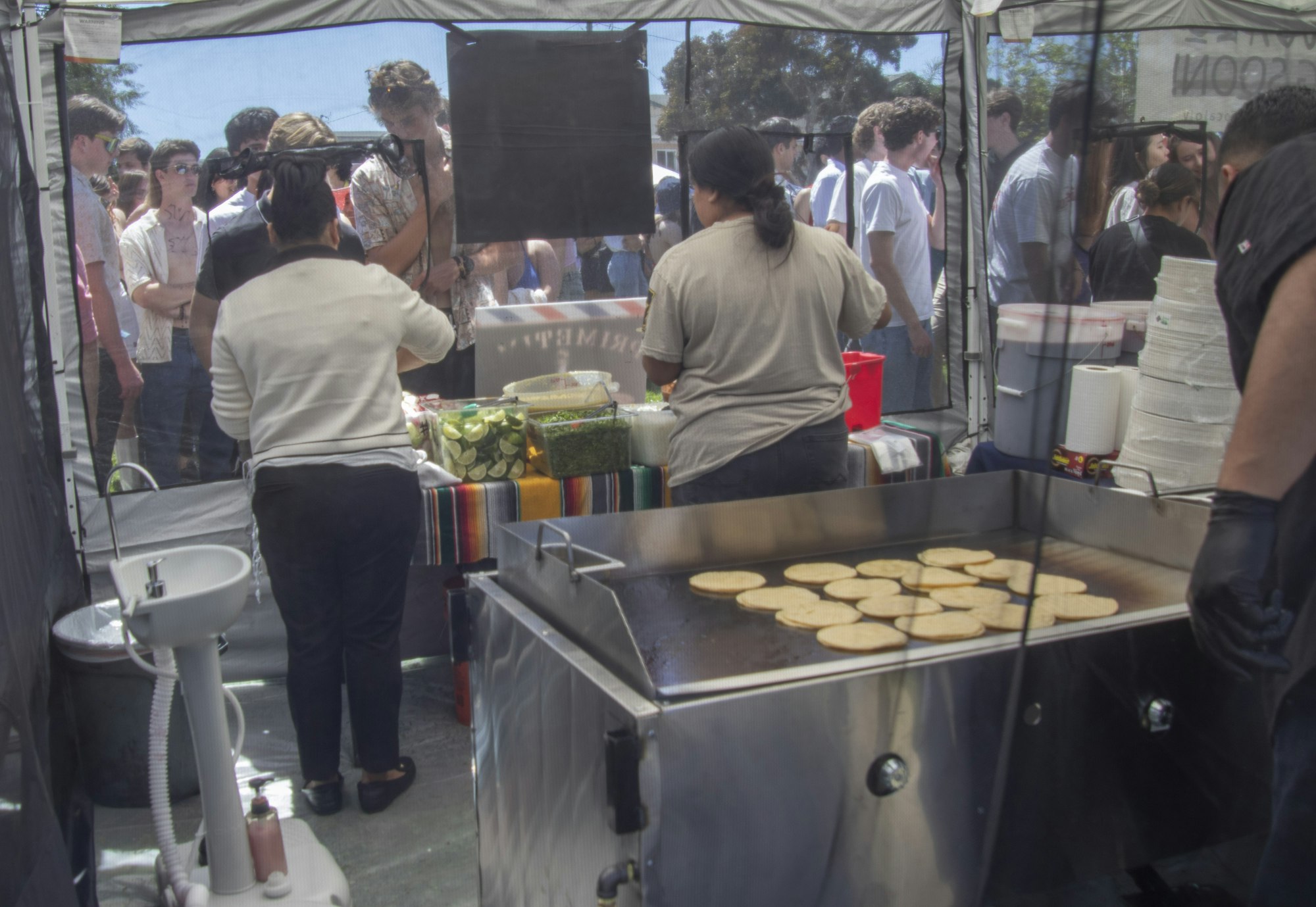 Outdoor food stand with workers and a queue of customers, cooking station in front with tortillas on the grill.