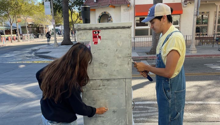 Two people working on a utility box on a sunny street with trees and buildings in the background.