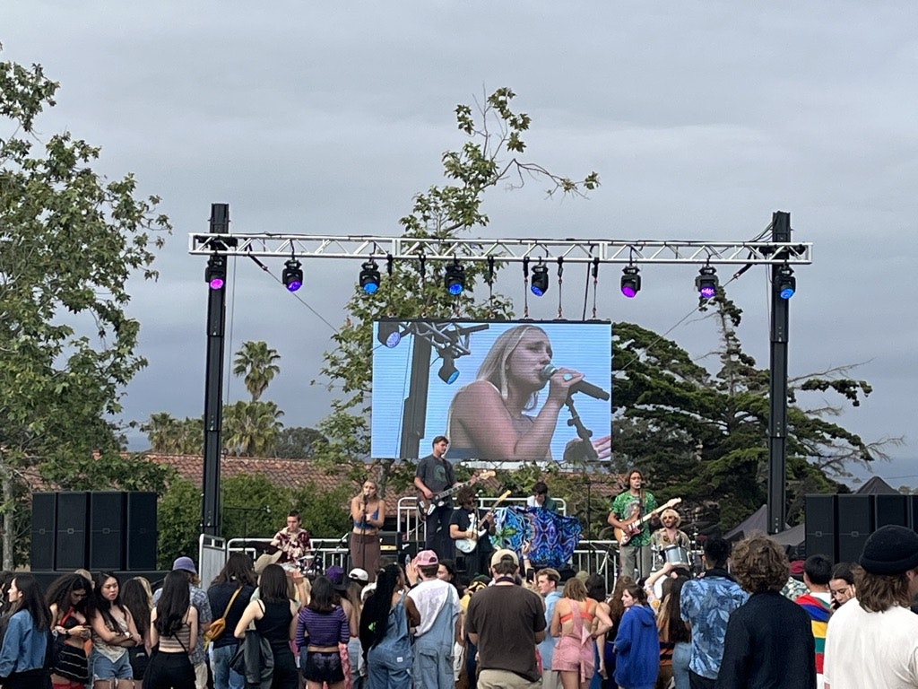 Outdoor concert with a crowd watching a band, stage lights, big screen showing a singer.