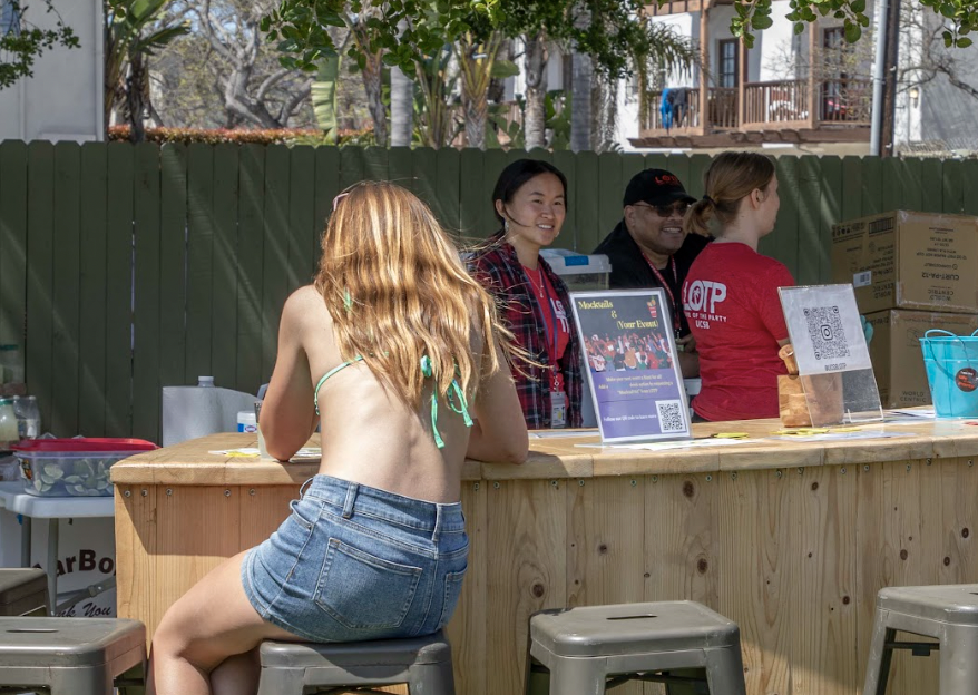 A sunny outdoor scene with people at a wooden bar, enjoying drinks and chatting, while someone sits at the bar.